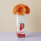 Photo du vase bouteille fraise avec des marguerites oranges. Il est posé sur une table lila devant un mur beige.