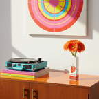 Photo du vase bouteille fraise mise en scène sur un meuble en bois brillant à côté d'un lecteur de vinyle et devant un tableau pop. Le vase contient un bouquet de marguerites oranges.
