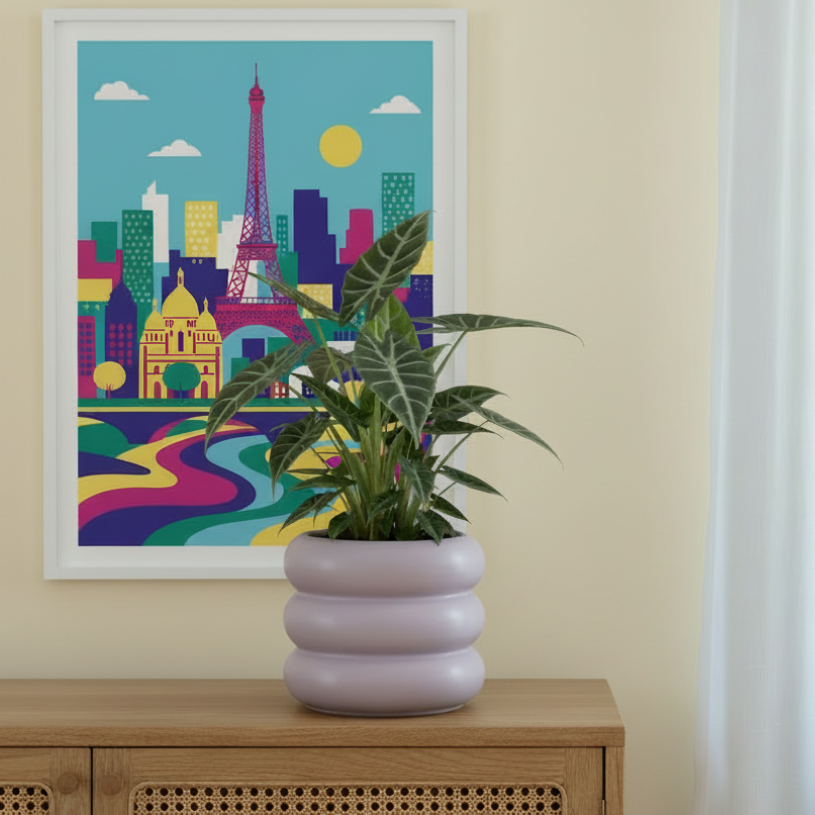 Photo de presentation du cache pot alocasia lila. Le cache pot est posé sur un meuble en bois avec du canage. Derrière il y a un tableau de paris. le mur est jaune clair.